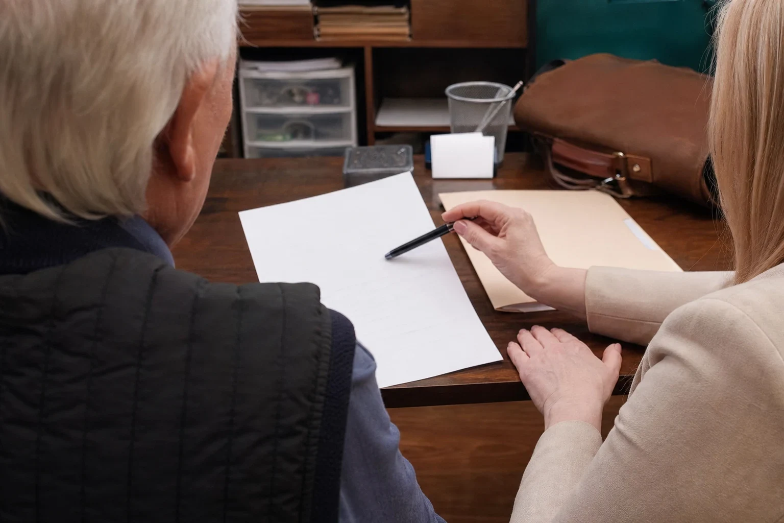 Over-the-shoulder view of an older man and a woman in a beige jacket at a wooden desk, her pen pointing to a sheet of paper beside a manila folder and leather bag